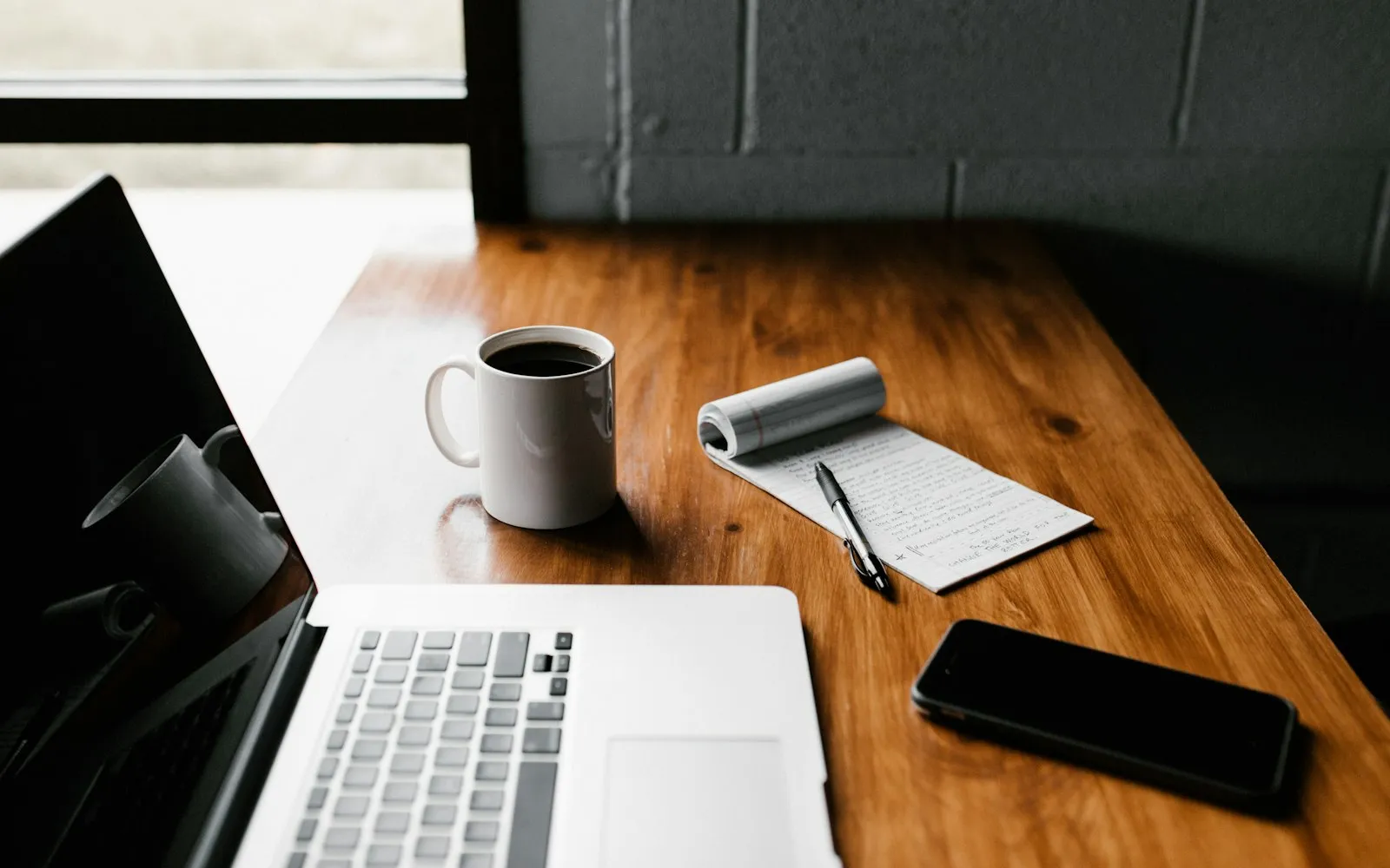 An open laptop, a white coffee mug, a notepad and a smartphone on a wooden desk by a window