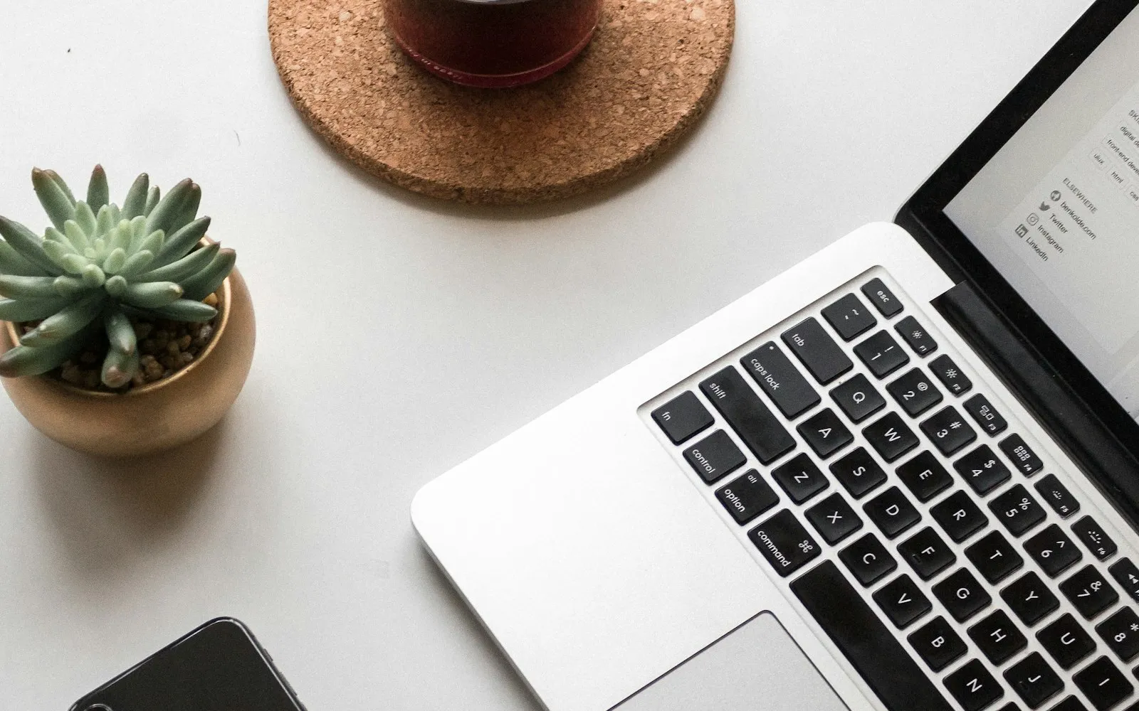 A smartphone, an open laptop, a small succulent and a drink on a white desk, seen from above