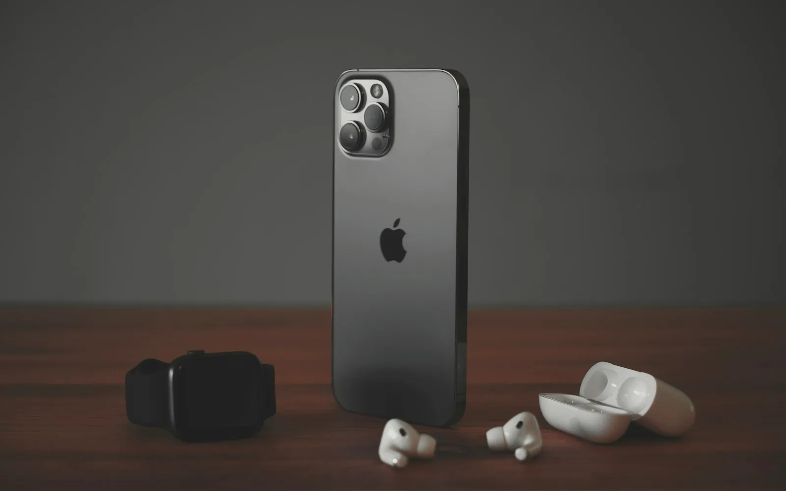 An iPhone standing on a wooden desk between an Apple Watch and a pair of wireless earbuds with their open case