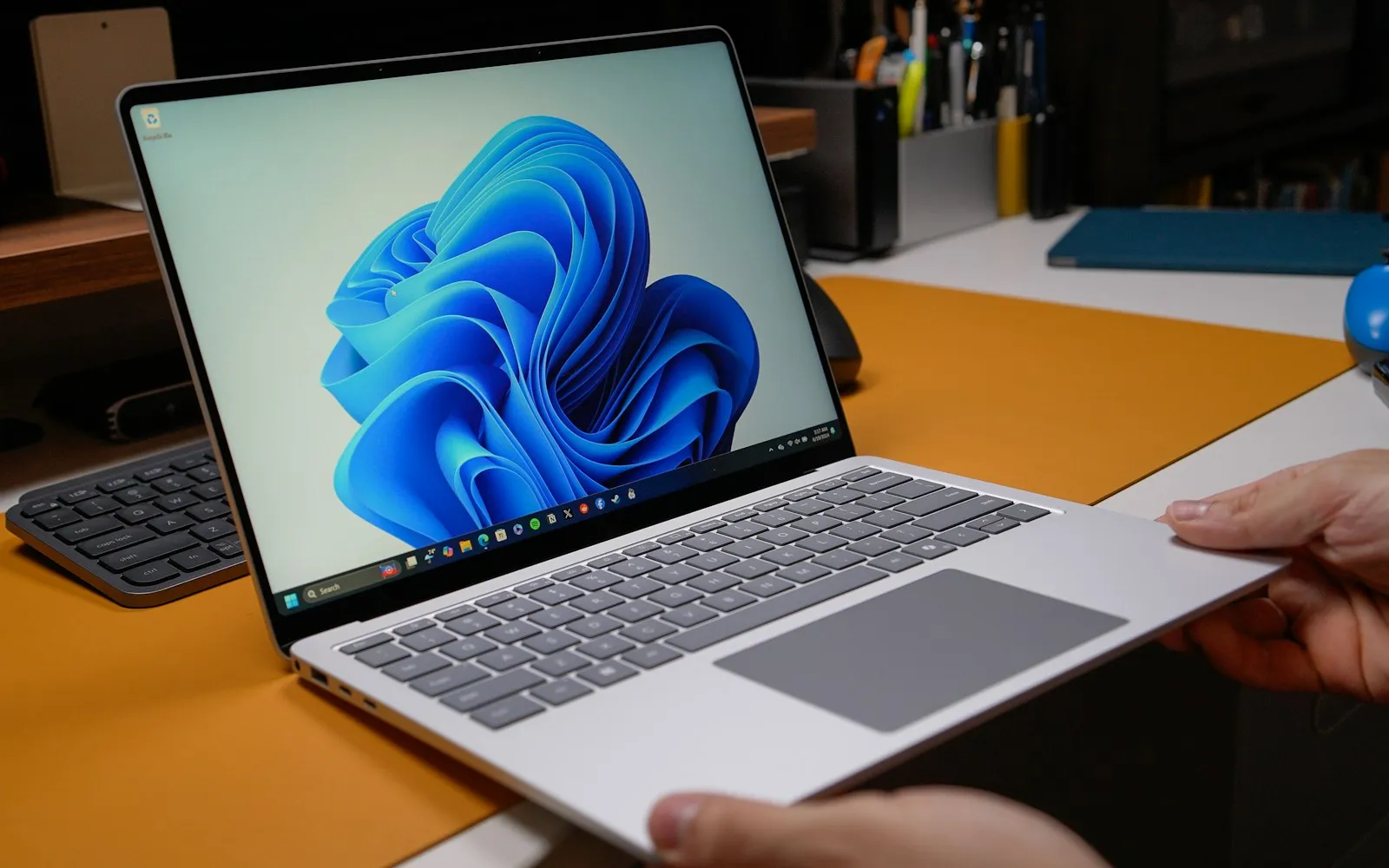 Hands holding an open Windows 11 laptop above a desk with an orange desk mat